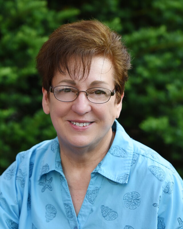 Color photograph of the smiling author, CJ Nicks, wearing a blue blouse and glasses.